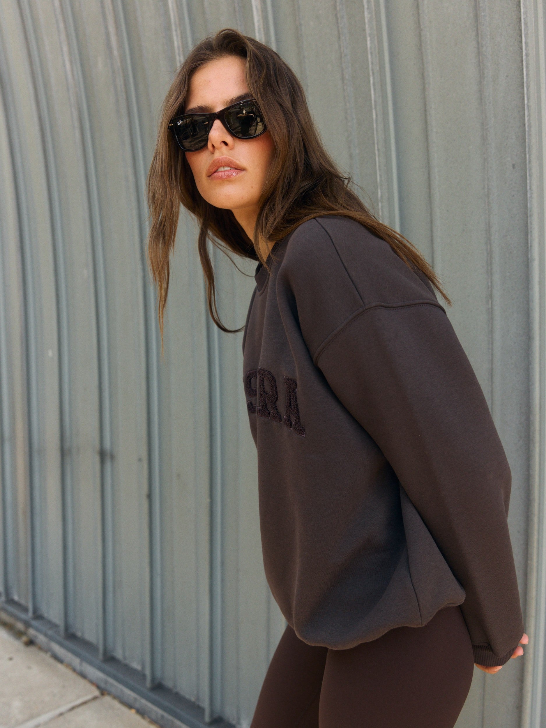 Woman wearing an espresso brown oversized crewneck sweatshirt against a ribbed metal wall.